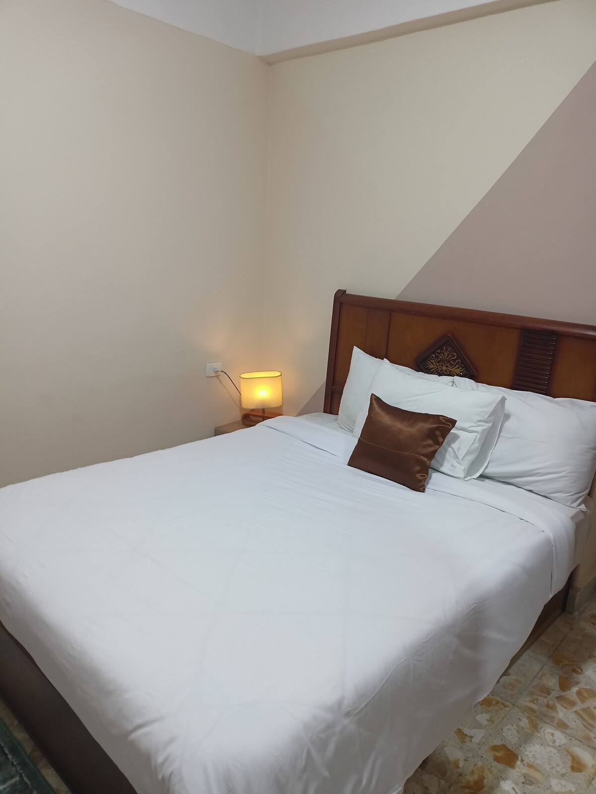 A cozy sleeping area features a neatly made bed with white linens and a decorative brown pillow. Soft lighting from a bedside lamp creates a warm glow, complementing the light-colored walls and wooden headboard.