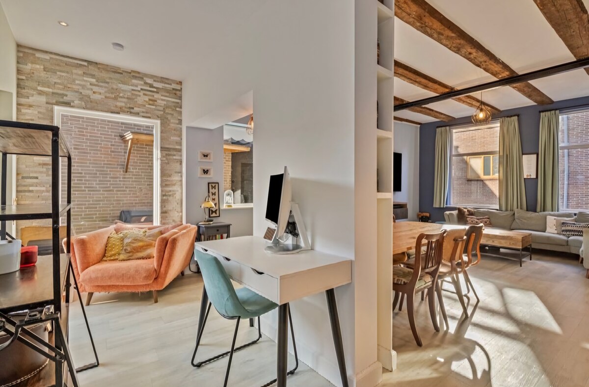 A spacious living area features a mix of modern and rustic design elements, including exposed wooden beams and a stone accent wall. A cozy lounge area contains an orange armchair, while a dining space shows wooden chairs arranged around a table, illuminated by natural light from large windows.