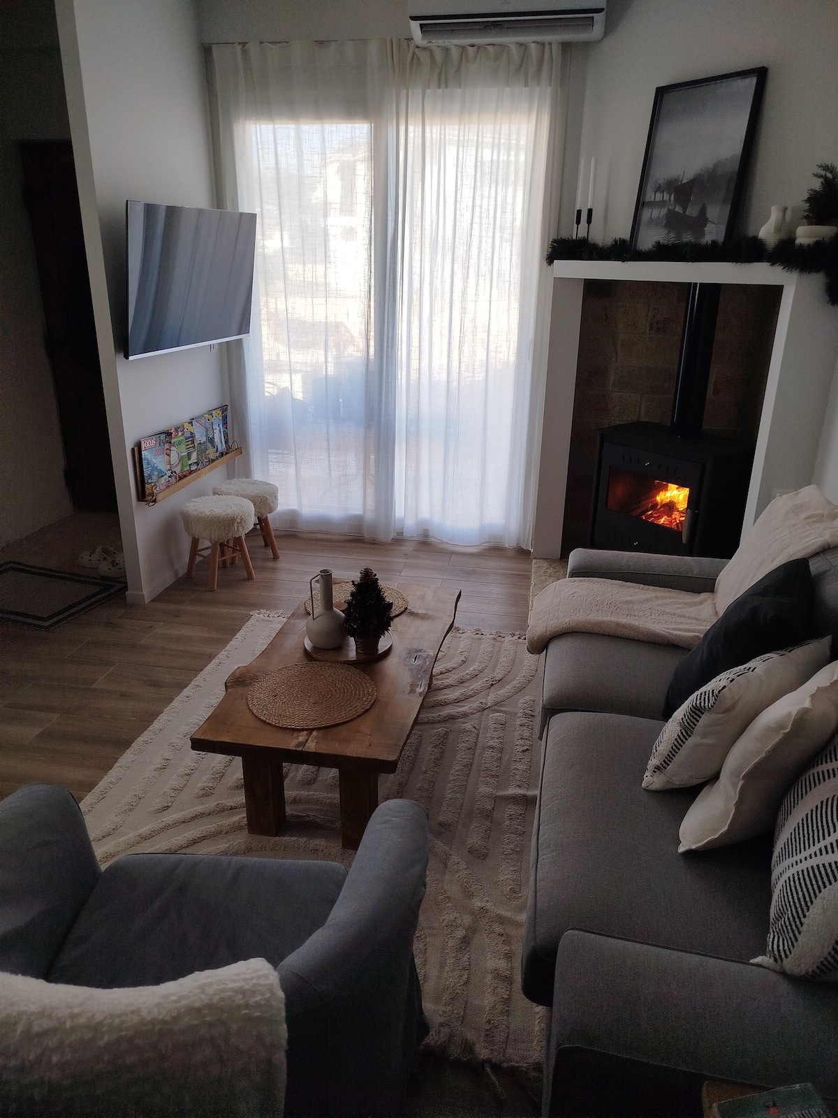 A cozy living area is furnished with a gray sofa and an accent chair. A wooden coffee table is centered on a light rug, while a softly glowing fireplace adds warmth. Large windows are draped with sheer curtains, allowing natural light to brighten the space.