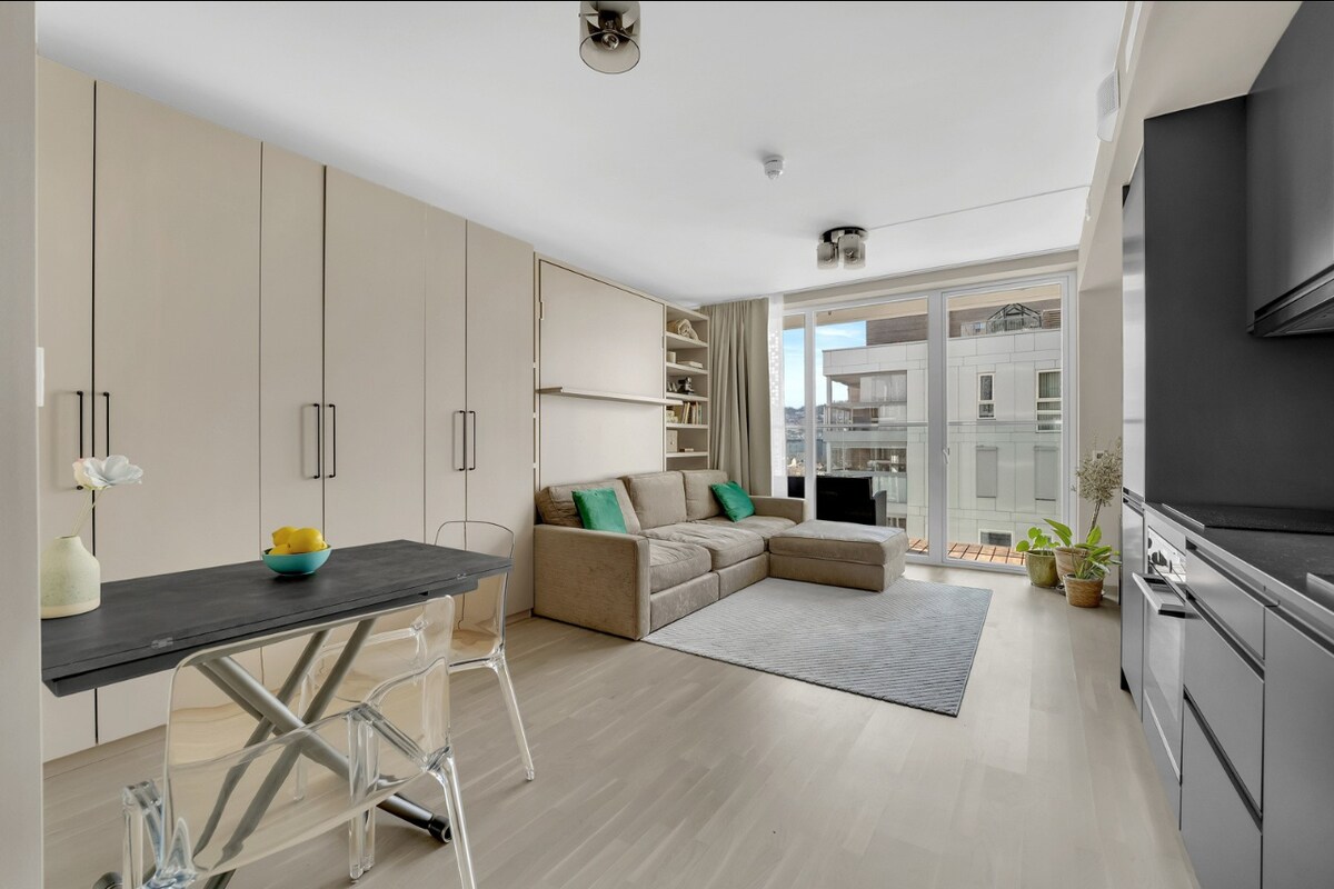 A bright and spacious living area is depicted, featuring a comfortable sofa with green accent cushions. A modern dining table is positioned nearby, complemented by clear chairs. Large windows provide an abundance of natural light and a view of the outdoors, enhancing the open feel of the room.