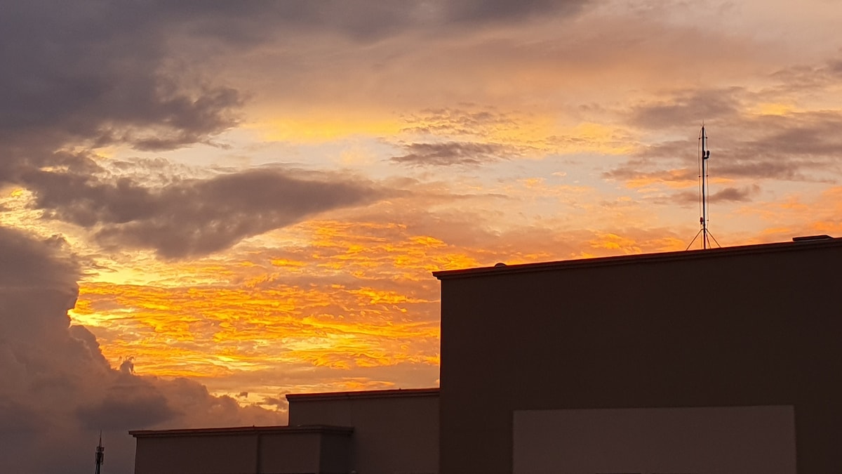 A vibrant sunset is captured with a palette of orange and yellow hues spreading across the sky. The silhouettes of rooftops are visible at the foreground, emphasizing the colorful sky above, while the outlines of structures add depth to the scene.