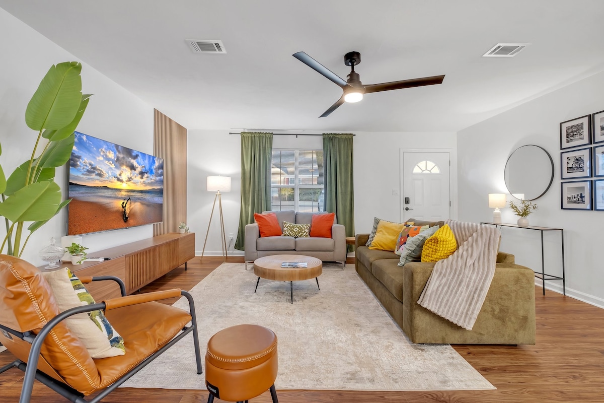 The living area features a spacious seating arrangement with a combination of a green couch, gray sofa, and leather armchairs. A large television is mounted on a wooden wall unit. Natural light enters through a window with green curtains, illuminating the stylish decor and warm wood flooring.