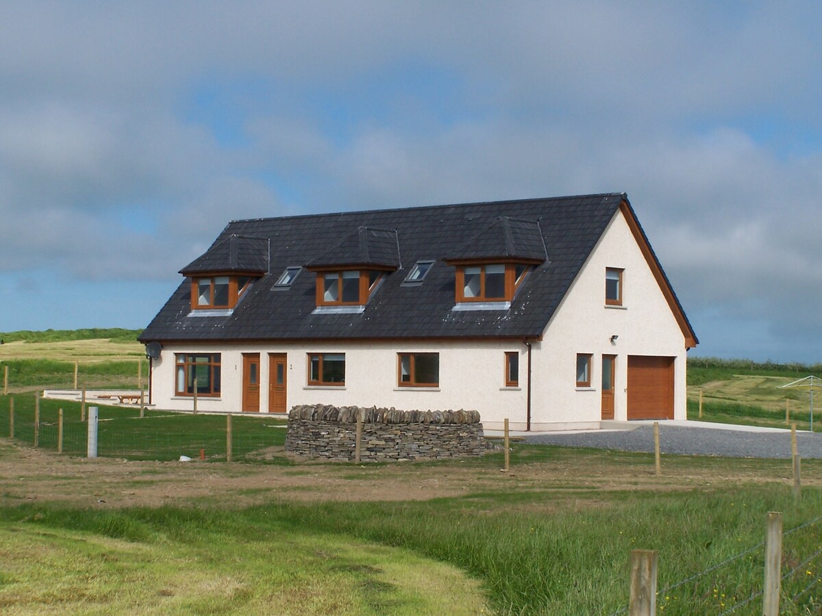 A two-story lodge is depicted, featuring a combination of windows and dormers under a slate roof. The exterior walls are painted in light colors. A stone wall is positioned in the foreground, with expansive green fields surrounding the structure.