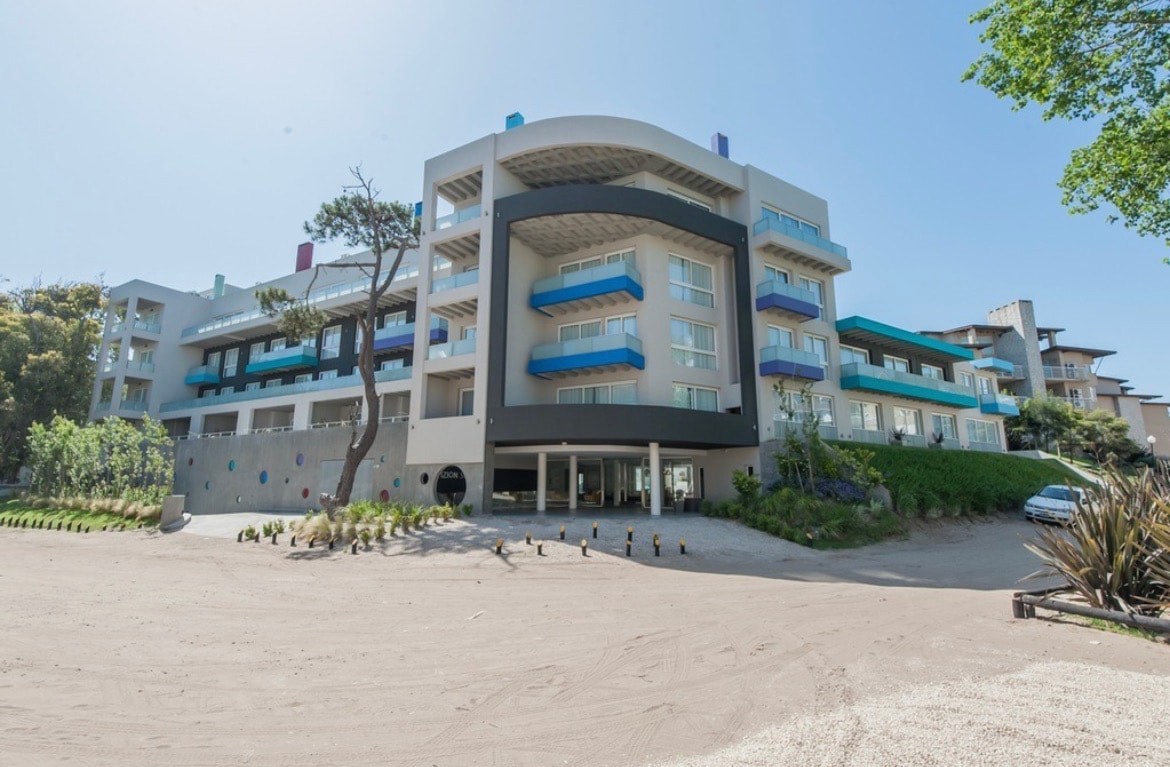 A modern, multi-story building is depicted, featuring large windows and balconies with colorful accents. Surrounding greenery is visible, along with a sandy entrance area. The structure appears well-maintained, with an organized layout highlighting its contemporary design.