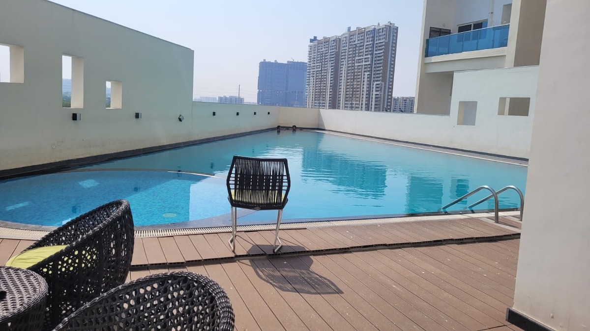 An outdoor pool area is highlighted, featuring clear blue water and a wooden deck. A single chair is positioned beside the pool, offering a space to relax. Modern buildings are visible in the background, with a bright sky adding to the atmosphere.