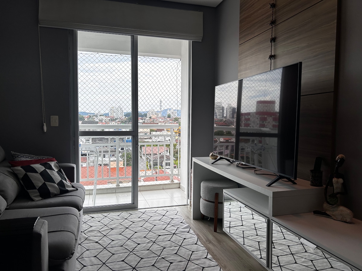 A modern living area is highlighted by a comfortable gray sofa and a large television on a minimalist white media console. Natural light floods the space through the glass sliding doors, which open to a view of the surrounding cityscape. A textured rug adds a cozy touch beneath the furniture.