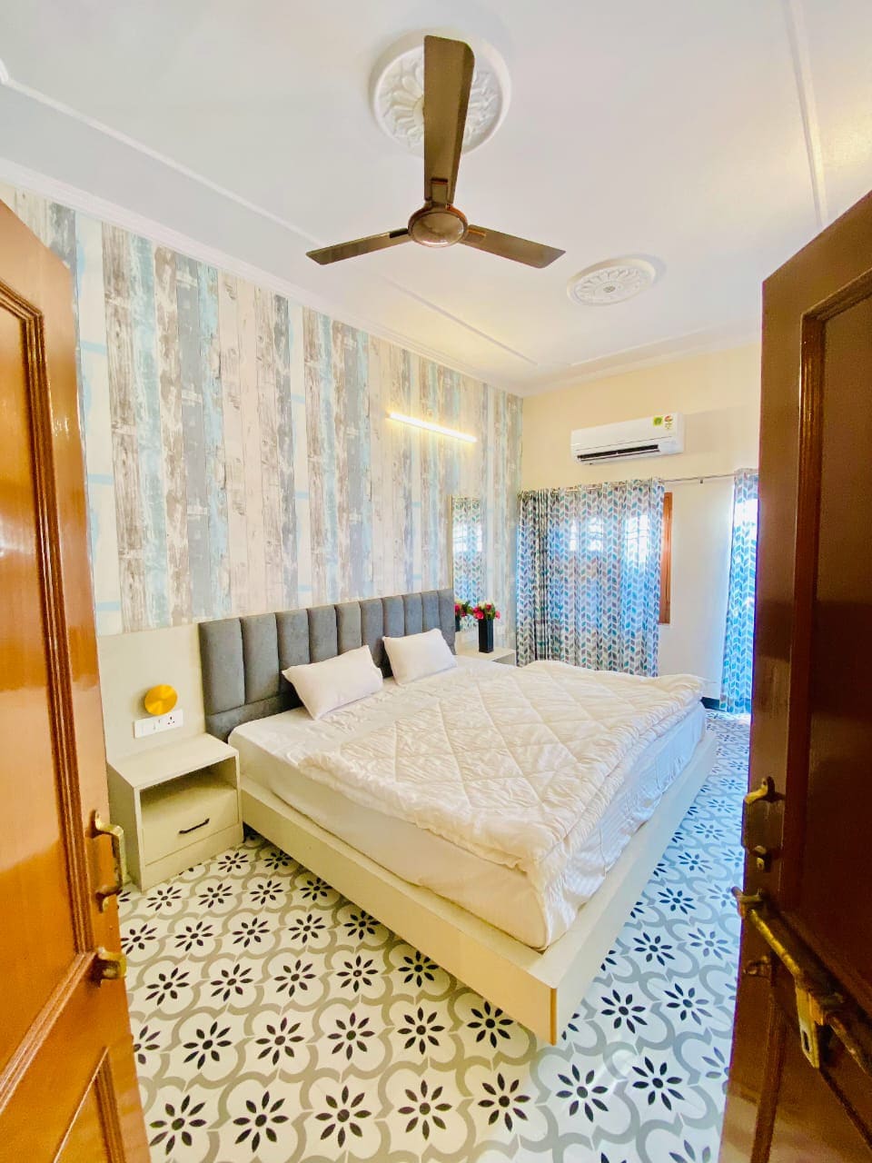 A cozy bedroom features a spacious bed with light linens, complemented by a stylish headboard. Decorative walls in muted tones and patterned tiles add character. Natural light filters through patterned curtains, and a small table displays a decorative item. An air conditioning unit is mounted for comfort.