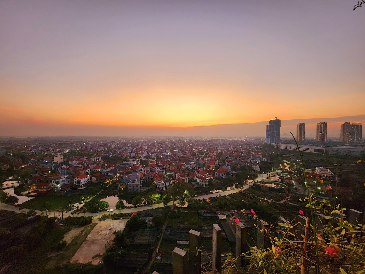 A panoramic view captures the blooming sunset over a residential area, with houses arranged in a vibrant patchwork. Waterways meander through the landscape, while distant buildings rise in the background, silhouetted against the evening sky.