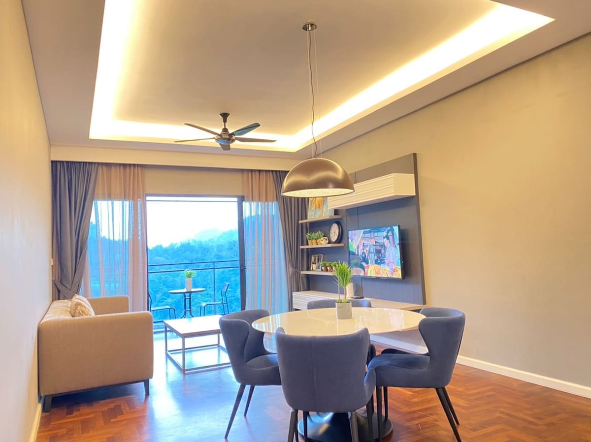 A modern living and dining area is highlighted by a round dining table surrounded by four upholstered chairs. A comfortable sofa is positioned to one side, with a large window providing natural light and access to a scenic outdoor balcony.