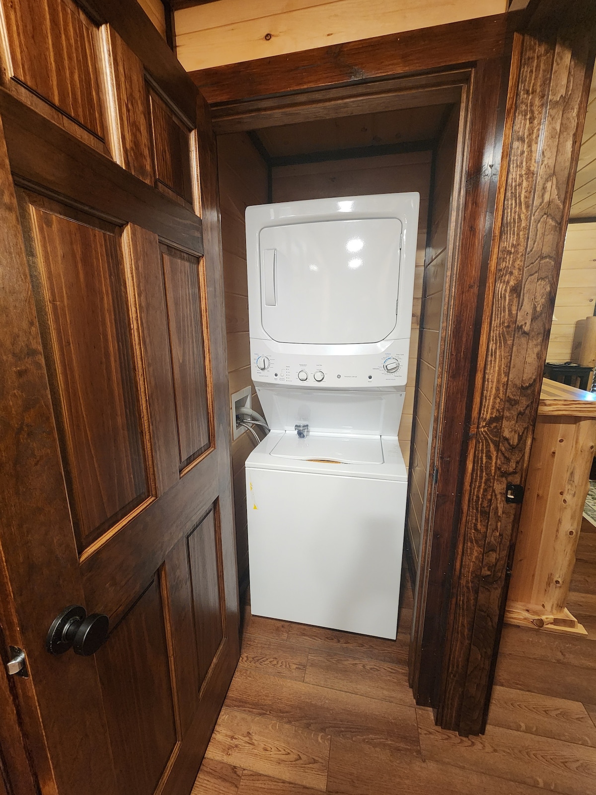 A stacked washer and dryer unit is positioned within a wooden closet, featuring a dark wood door with a simple handle. The surrounding walls are lined with light wood paneling, offering a warm and natural ambiance.