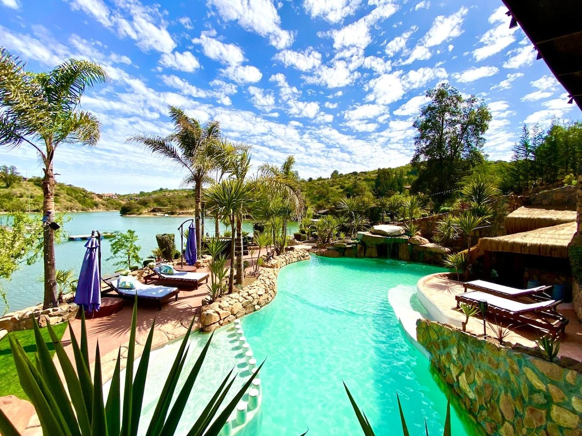 A tranquil pool area is surrounded by lush greenery and palm trees, with lounge chairs positioned along the water's edge. The pool features a gentle curve and transitions from light to deep turquoise, harmonizing with the natural landscape and clear blue sky dotted with clouds.