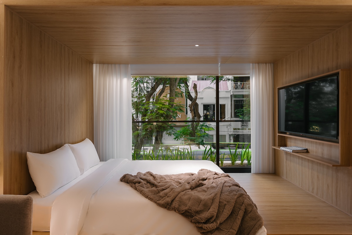 A modern sleeping area features a custom-designed king-sized bed with crisp white linens and a cozy throw blanket. Large floor-to-ceiling windows allow natural light to fill the space, offering views of lush greenery and the street outside. A flat-screen smart TV is mounted on the wall.