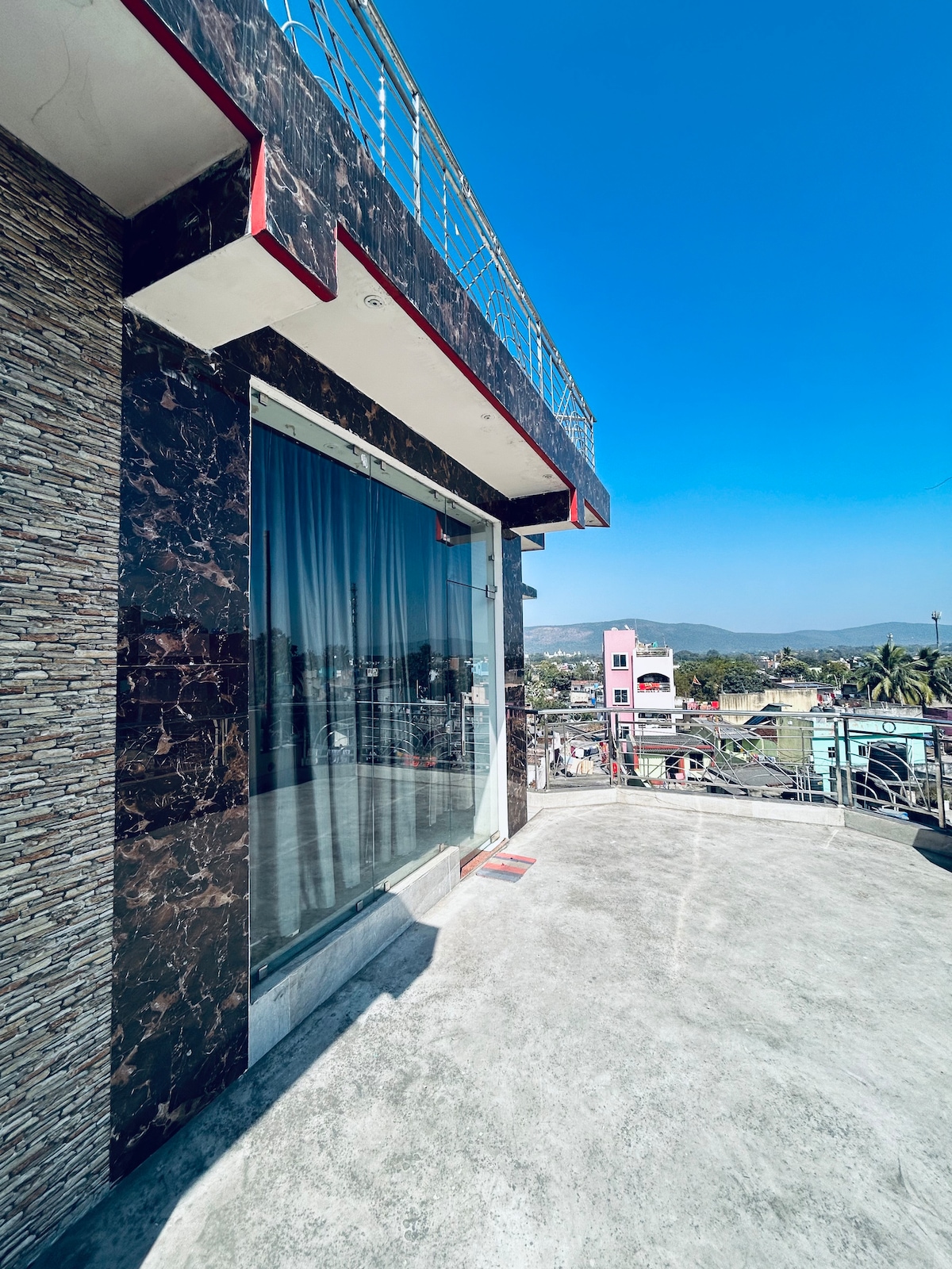 A spacious balcony is shown with a polished concrete floor and a large glass sliding door, offering views of the surrounding neighborhood. The area is framed by a decorative stone wall and metal railings, with a clear blue sky in the background.