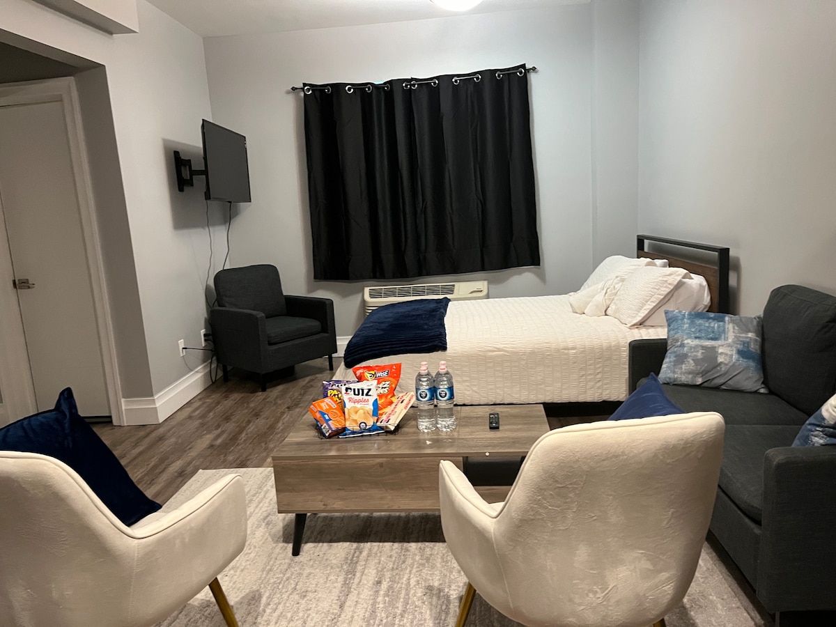 A furnished living space displays a spacious bed with crisp bedding. Two plush chairs and a gray sofa are arranged around a central coffee table, which holds snacks and bottles of water. A wall-mounted television is positioned in front of the seating area, and dark curtains cover the window.