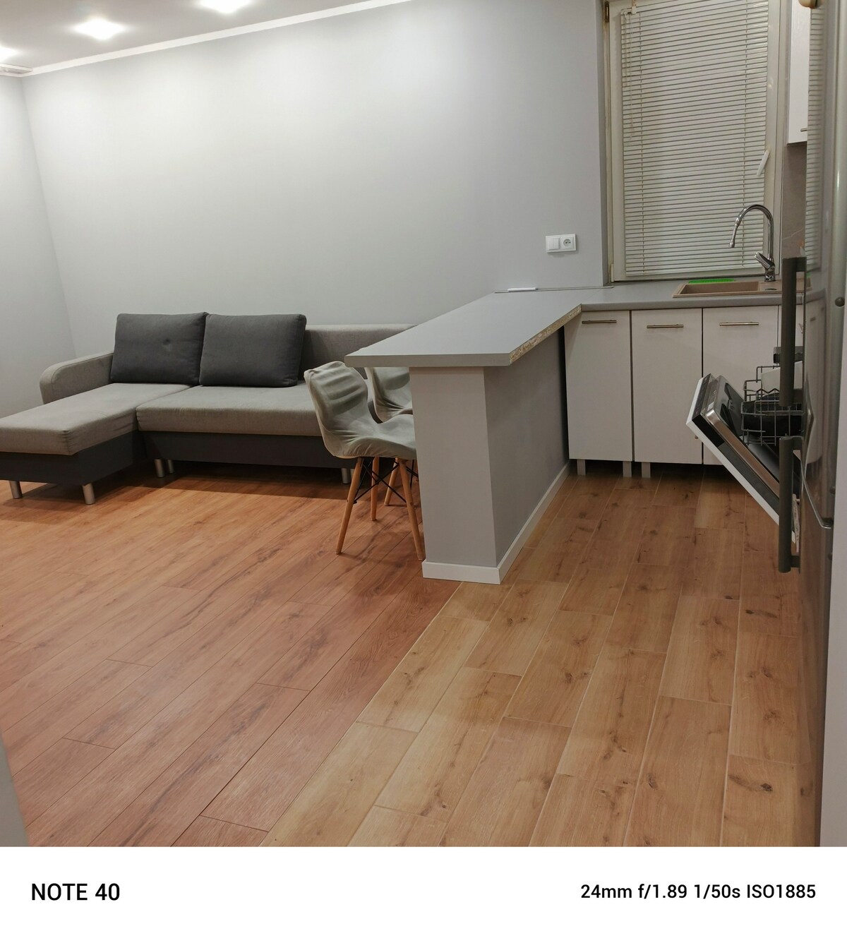 A modern living area features a soft gray sectional sofa positioned beside a minimalistic kitchen bar. Light wood flooring adds warmth to the space, while a large window allows for natural light. Kitchen appliances are neatly arranged, enhancing the functionality of the area.