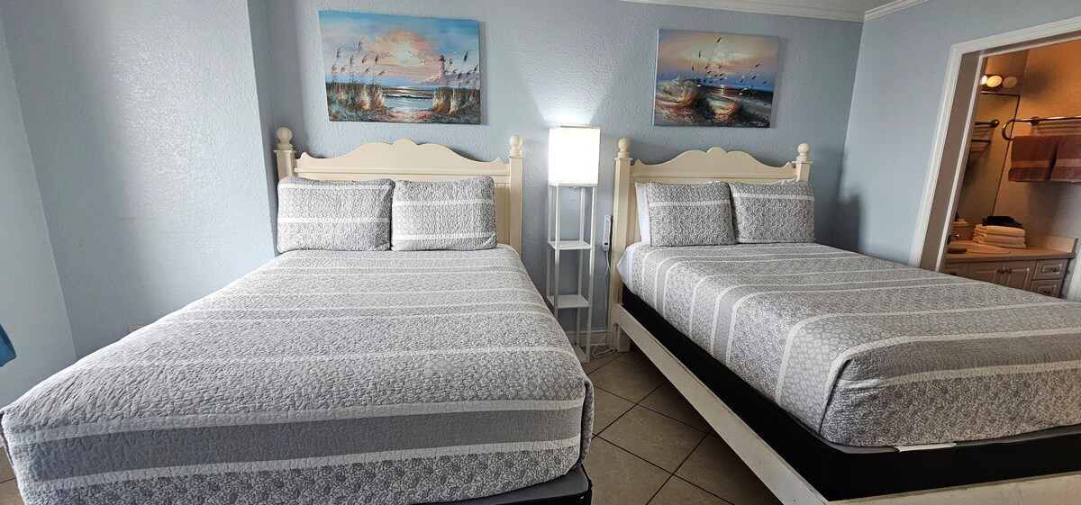 A brightly lit sleeping area is furnished with two large beds featuring decorative bedding. Art depicting serene coastal scenes hangs above the beds. A floor lamp provides additional light, and an open bathroom can be seen in the background, enhancing the overall functionality of the space.