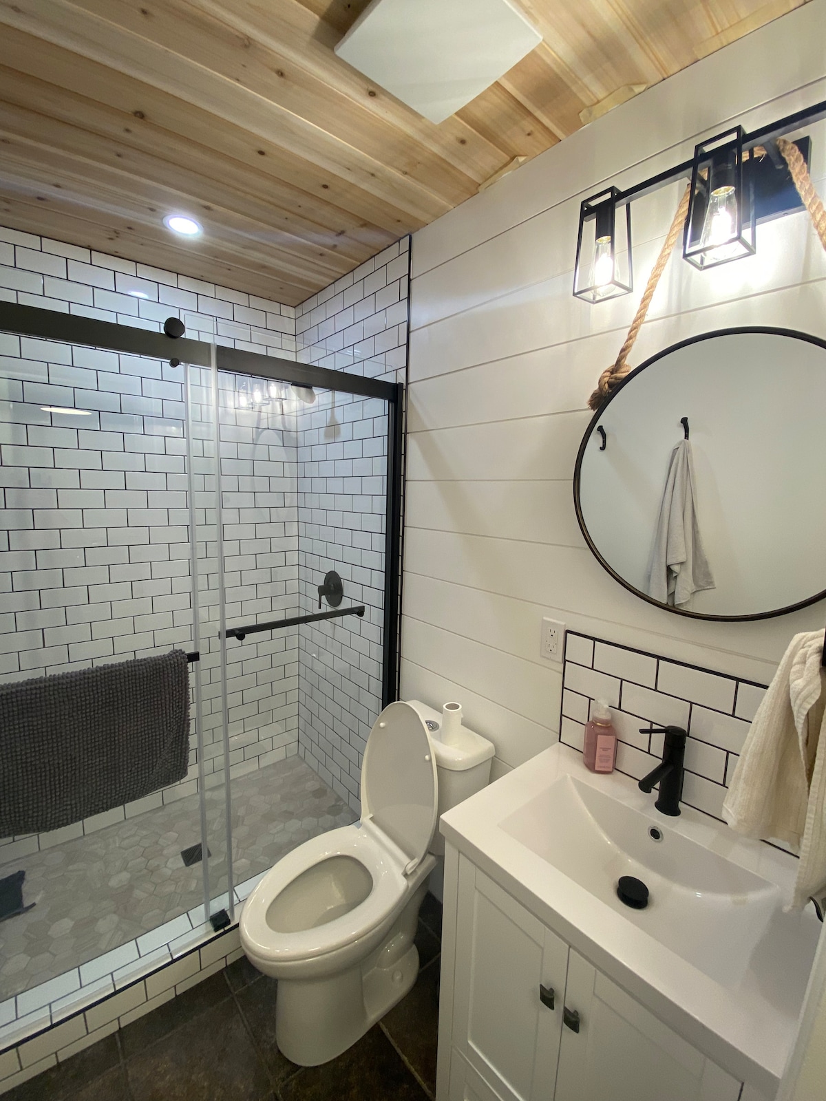 A modern bathroom is showcased, featuring a glass shower enclosure with white subway tile. A round mirror is mounted above a vanity with a basin, while a towel hangs from a hook nearby. The ceiling displays wooden panels, adding warmth to the space.