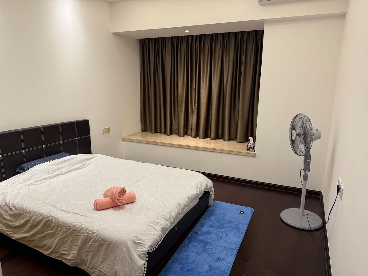 A spacious master room is presented, featuring a large bed adorned with a light bedspread. A pink towel is placed neatly on the bed. A fan stands on the floor next to a window adorned with dark curtains, allowing soft light to filter into the room.