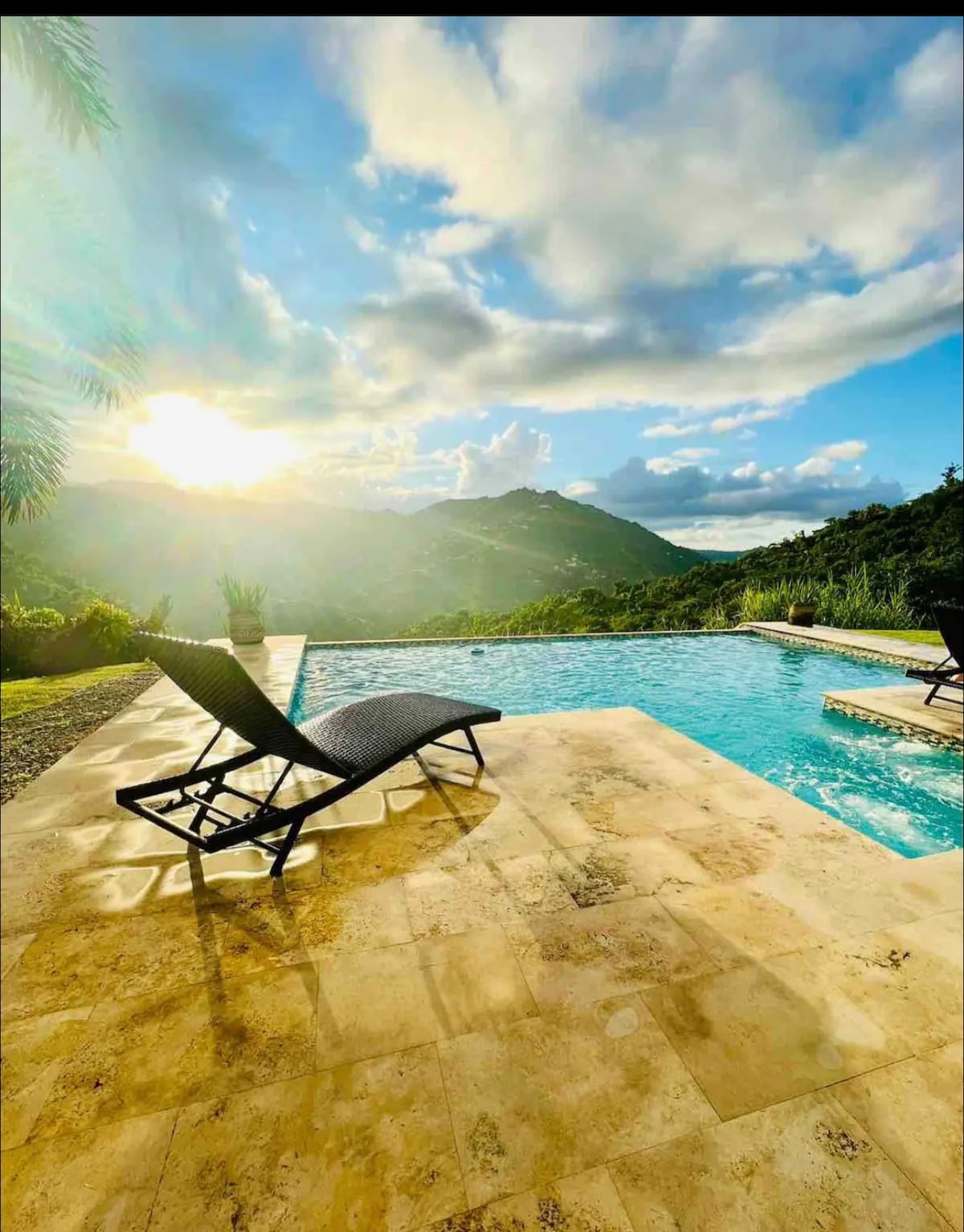An infinity pool is visible, overlooking lush hills. Sunlight glimmers on the water's surface, creating a tranquil atmosphere. A sun lounger with a black frame is positioned next to the pool, inviting relaxation amidst the natural surroundings.