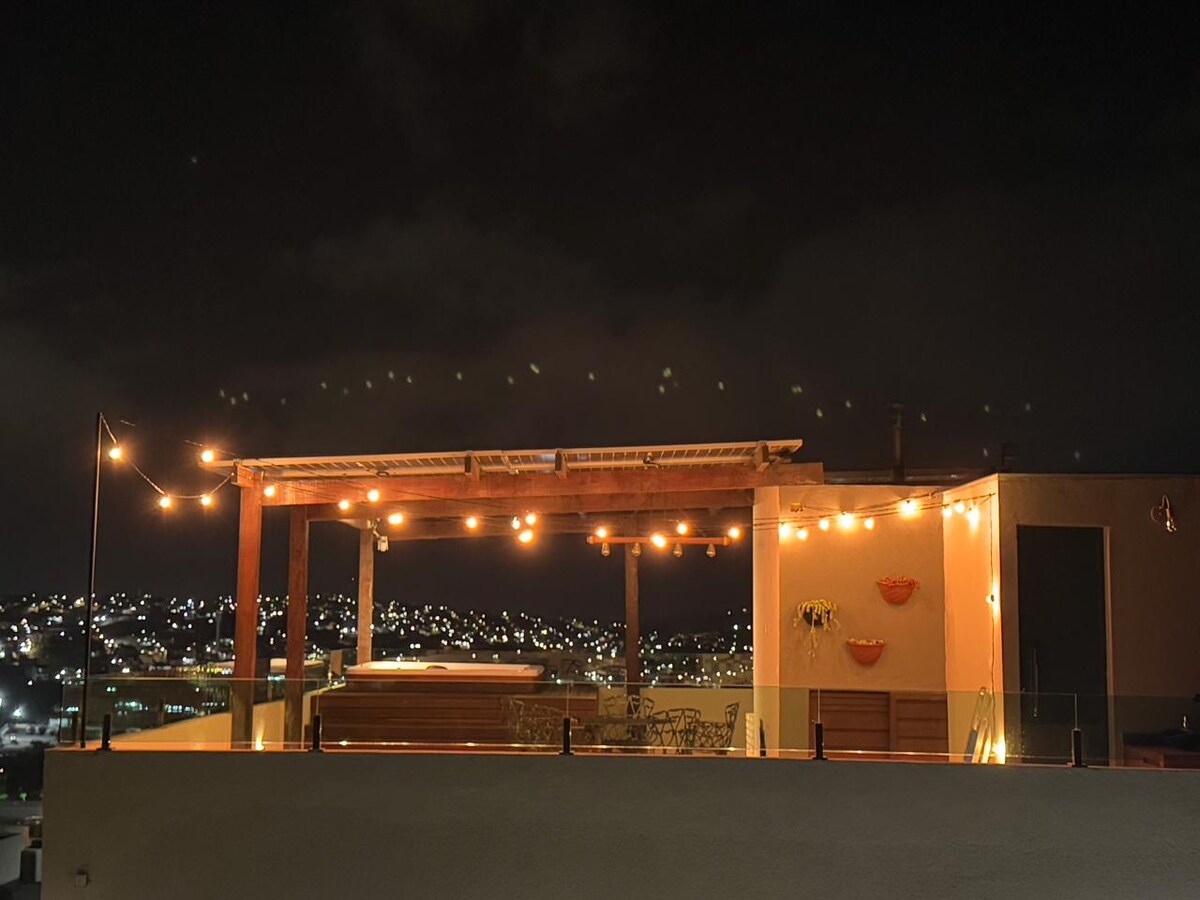 A rooftop area is illuminated by warm string lights, featuring a cozy seating arrangement. A Jacuzzi is positioned against a backdrop of twinkling city lights, creating a serene ambiance. The space is enclosed with glass railings, enhancing the unobstructed views of the surroundings.