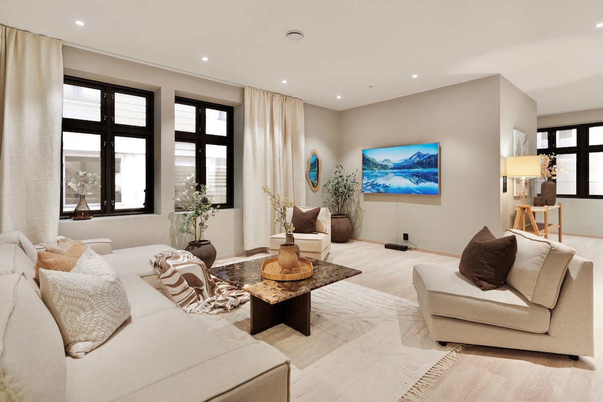 A spacious living area is highlighted by large windows that provide natural light. A comfortable sectional sofa is arranged around a coffee table, with minimalistic decor, including plants and wall art. A large television is mounted on the wall, offering recreational options.