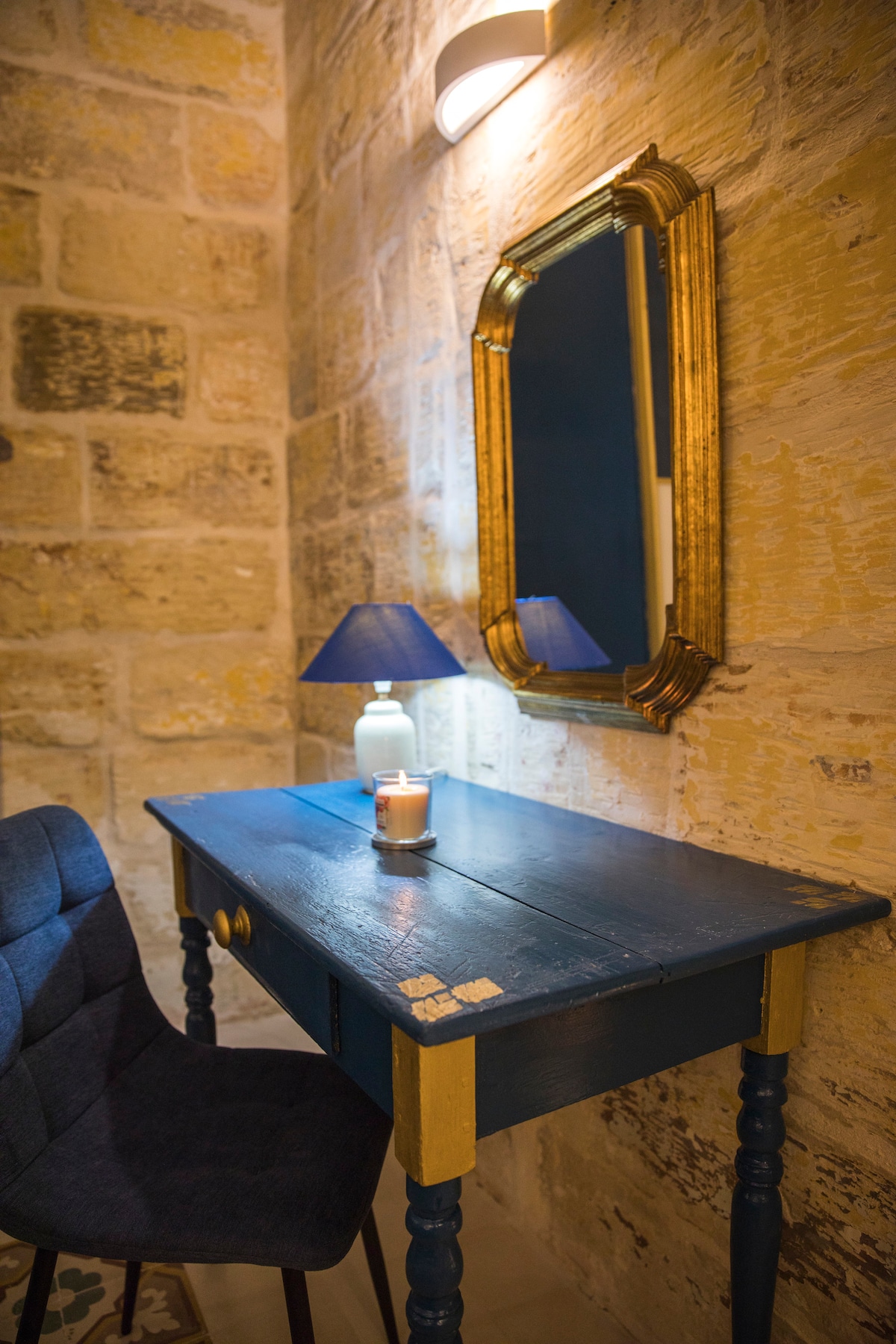 A wooden desk in a deep blue tone is paired with a dark upholstered chair. A small lamp with a blue shade sits on the desk, next to a candle. A decorative mirror with a gold frame is mounted on the stone wall, enhancing the area's unique charm.