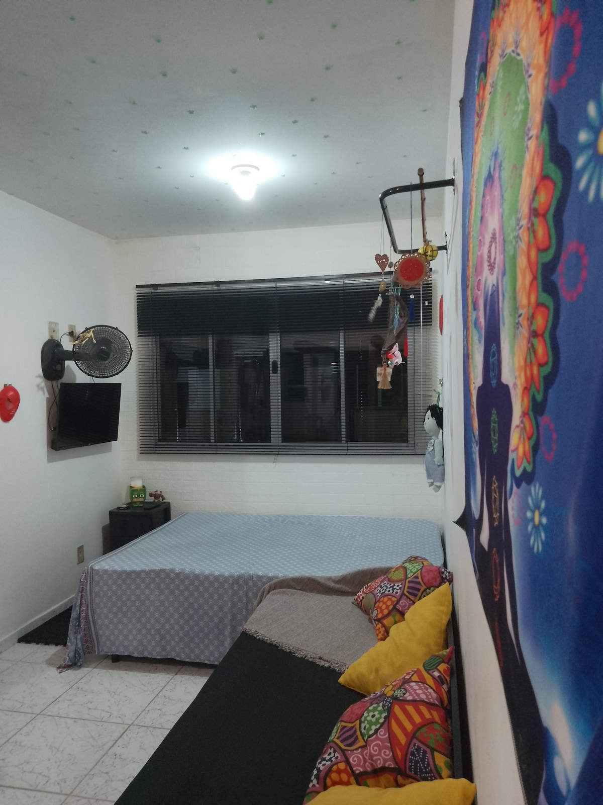 A cozy room features a spacious bed covered with a light blue quilt, complemented by decorative pillows on a black sofa. Natural light enters through a large window with horizontal blinds, and a colorful wall hanging adds a vibrant touch to the atmosphere.