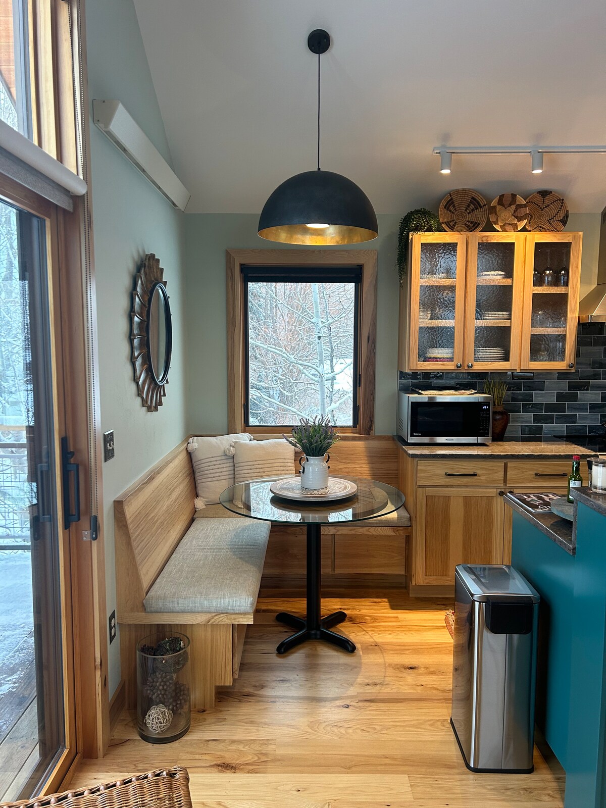 A corner dining area features a circular glass table surrounded by a wooden bench and a chair. Natural light filters through a window adorned with a simple curtain. A black pendant light hangs above, and wooden cabinetry is seen in the background, housing kitchen appliances.