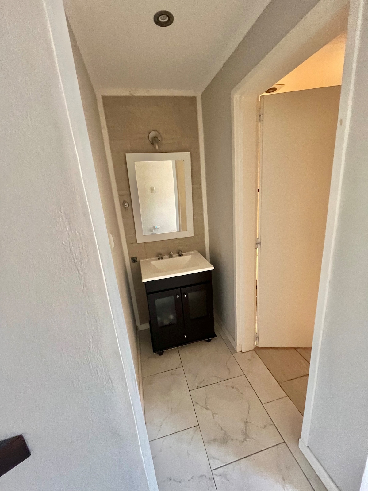 A compact bathroom area features a modern sink atop a dark cabinet, with a large mirror above it. The walls are adorned with light tiles, and soft lighting is provided by ceiling fixtures. An open door leads to a separate space beyond.