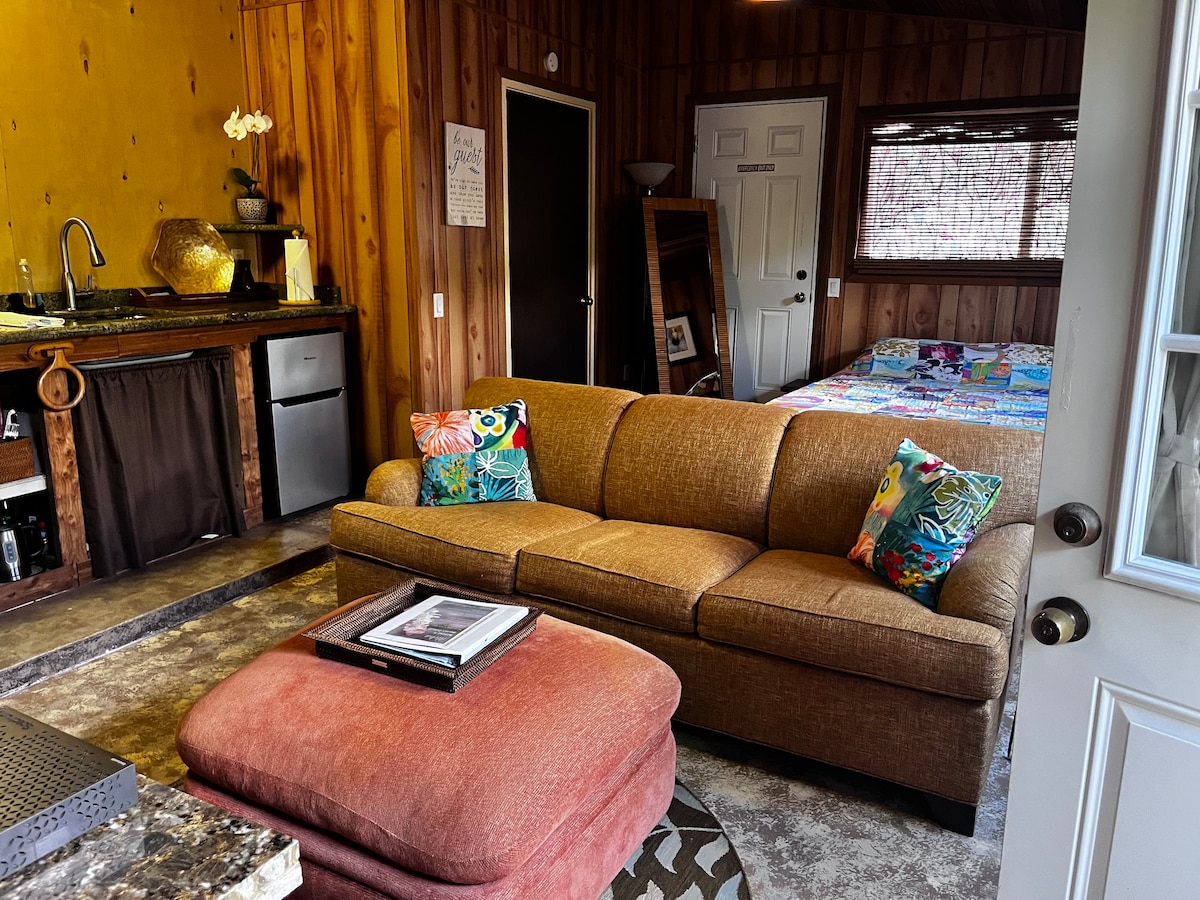 A cozy living area features a comfortable sofa with decorative pillows, paired with an ottoman. A kitchenette is visible in the background, displaying a sink and mini fridge. A patterned quilt covers the bed, partially seen in the adjacent sleeping area.