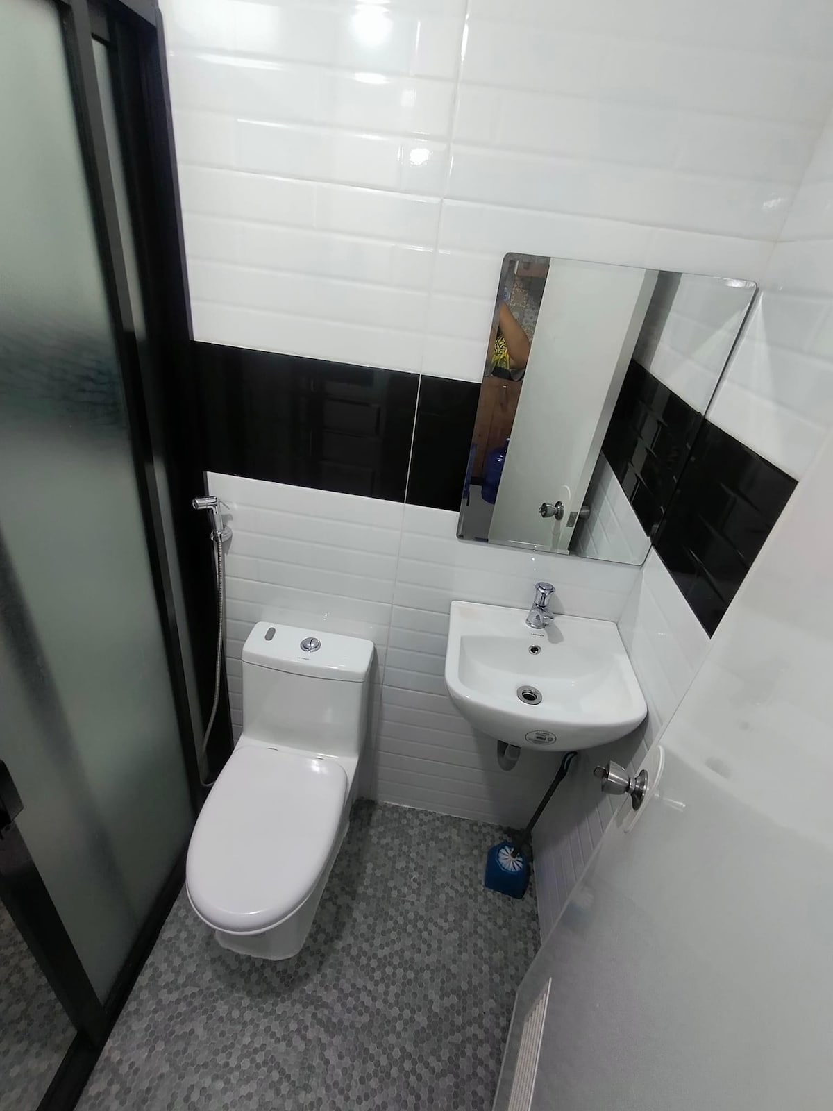 A modern bathroom features a shower enclosed in a glass panel, a wall-mounted mirror above a compact sink, and a sleek toilet. The space is elegantly tiled with a combination of glossy white and black tiles, creating a clean and functional appearance.