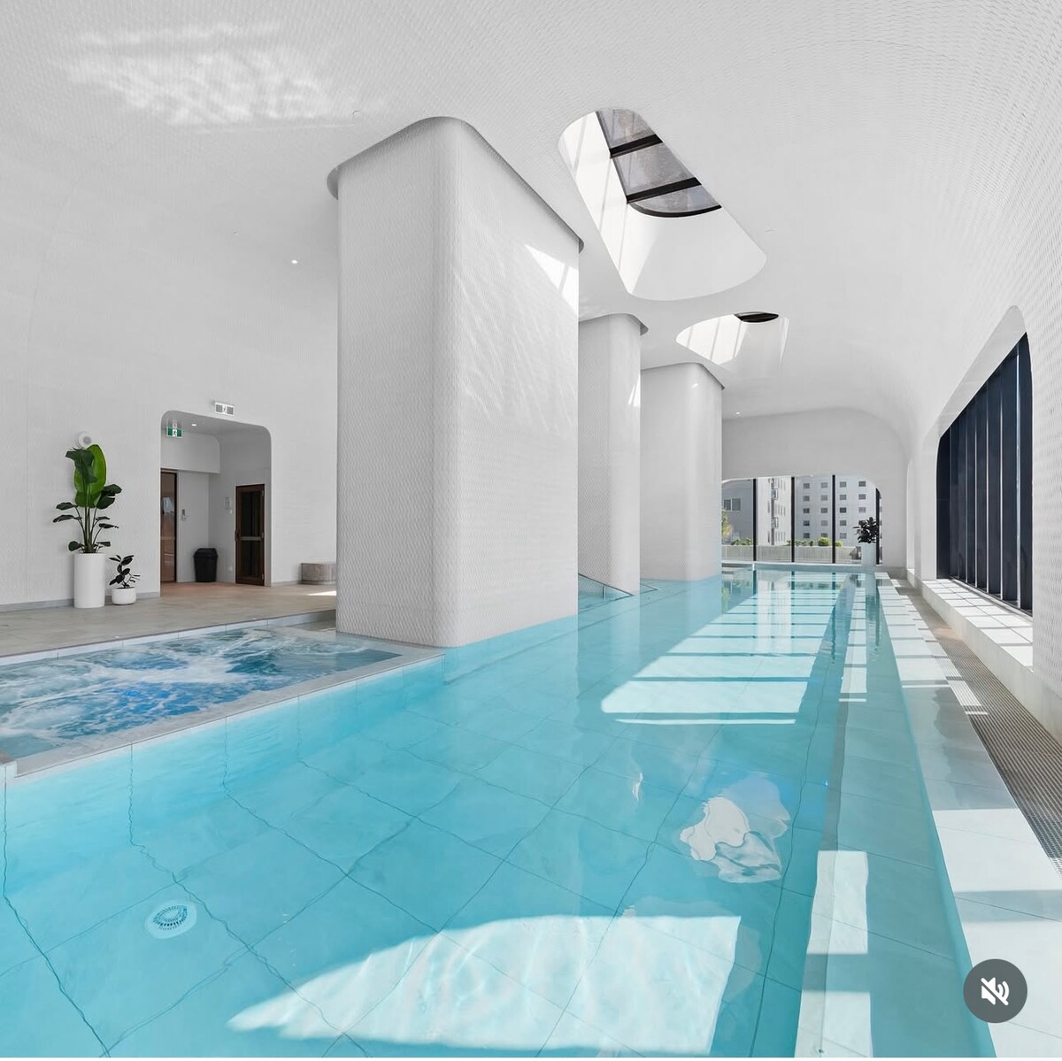 A spacious indoor pool area features clear turquoise water reflecting light from skylights above. Tall, white walls create an airy atmosphere, complemented by strategic plant placements. Large floor-to-ceiling windows allow for natural light, enhancing the serene environment.