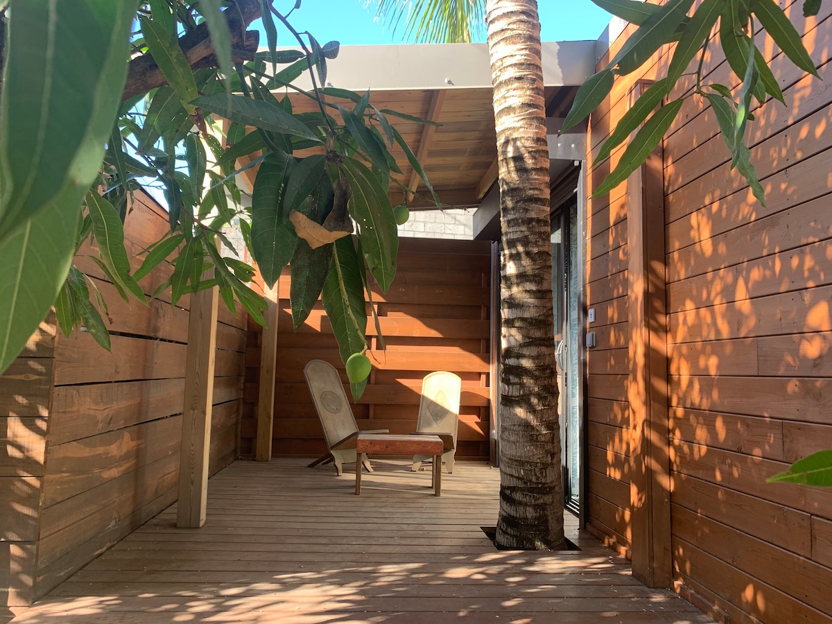 An outdoor seating area is surrounded by wooden walls, featuring two lounge chairs and a small table. Sunlight filters through foliage, casting playful shadows on the wooden deck. The space is complemented by a palm tree, adding a natural touch to the environment.
