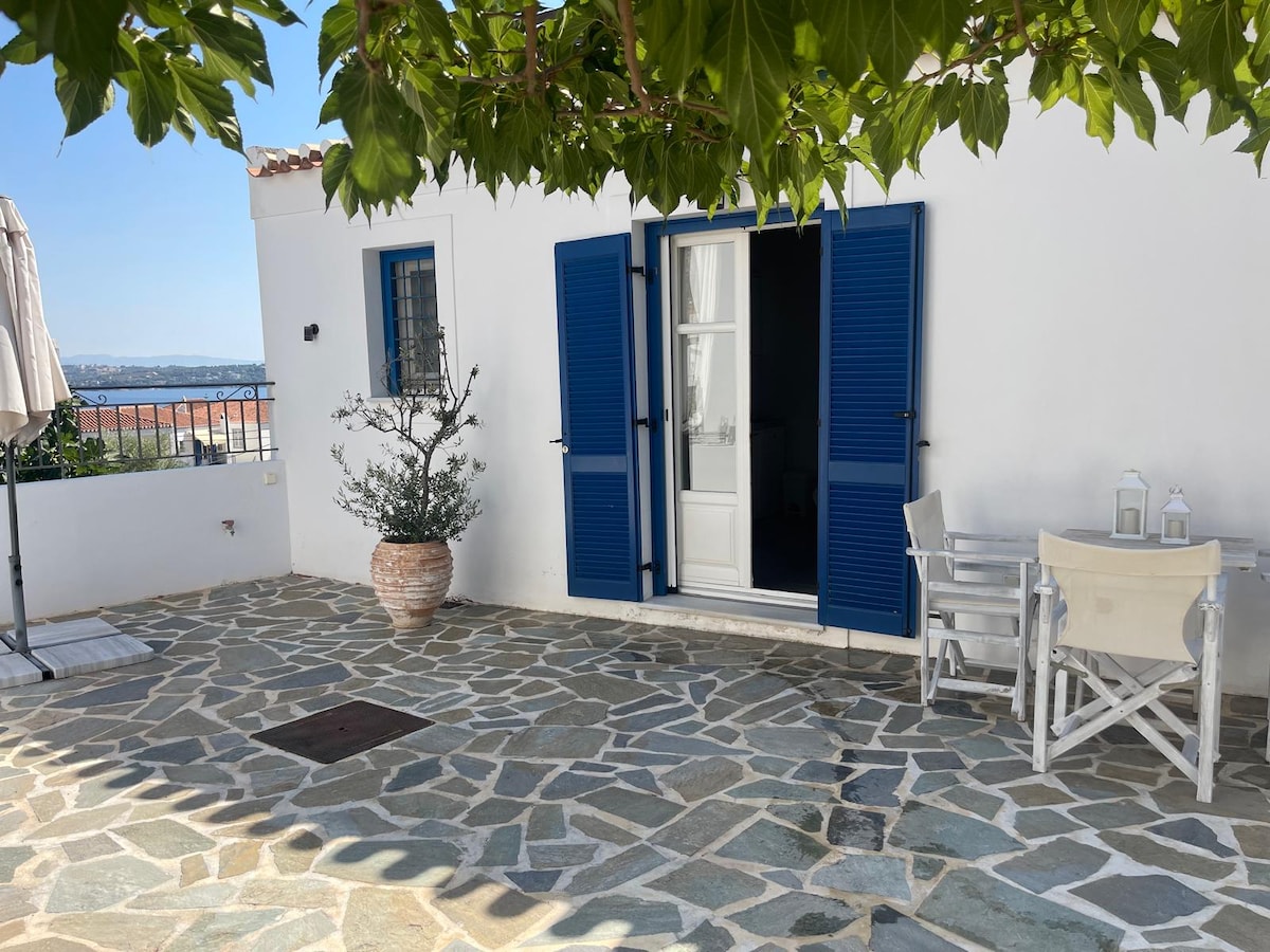 A charming outdoor courtyard features a stone floor, framed by a lush green tree and a decorative pot. Blue wooden shutters open to reveal a white door, accompanied by a small table and two chairs, ideal for enjoying the serene setting.