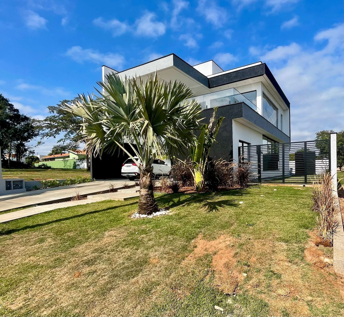 A modern two-story house is set against a clear blue sky, surrounded by well-maintained greenery. Palm trees and ornamental plants adorn the front yard, providing a lush contrast to the sleek architecture. A driveway leads to a covered parking area on the left.