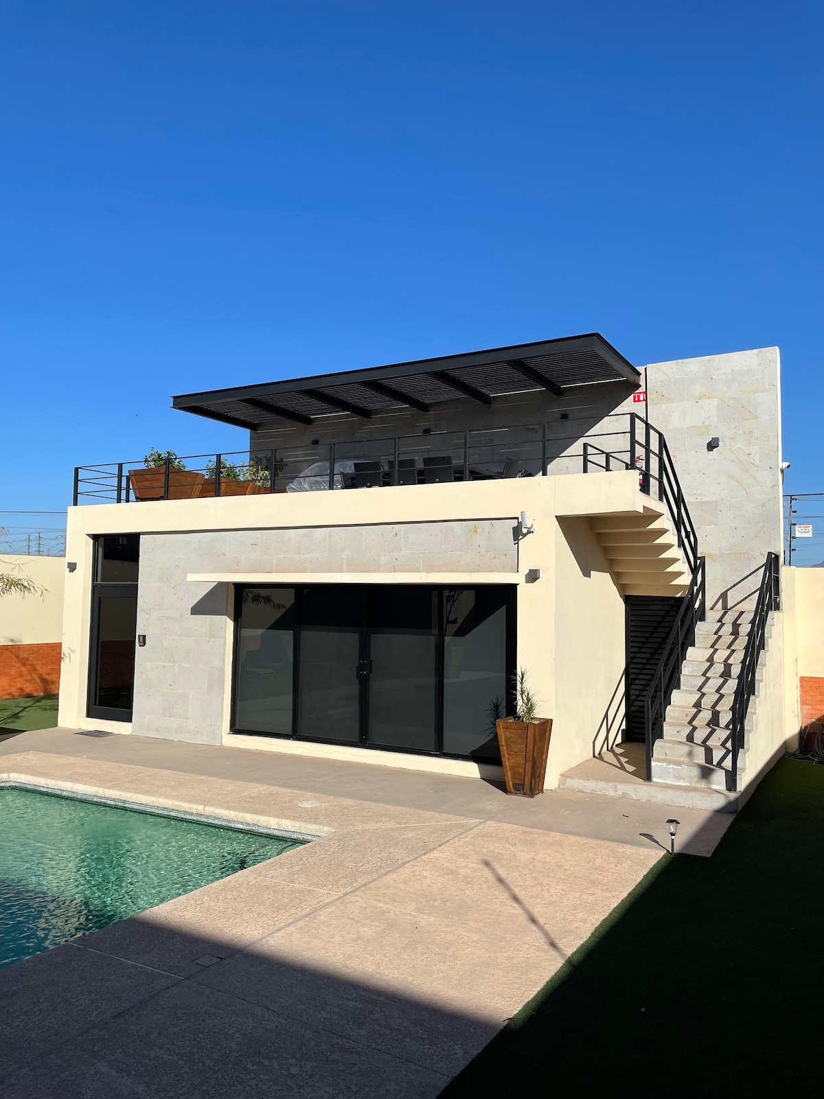 The exterior of a two-story residence is showcased, featuring large glass doors and a staircase leading to a rooftop area. A pool is visible in the foreground, and the property is surrounded by greenery and a clear blue sky.