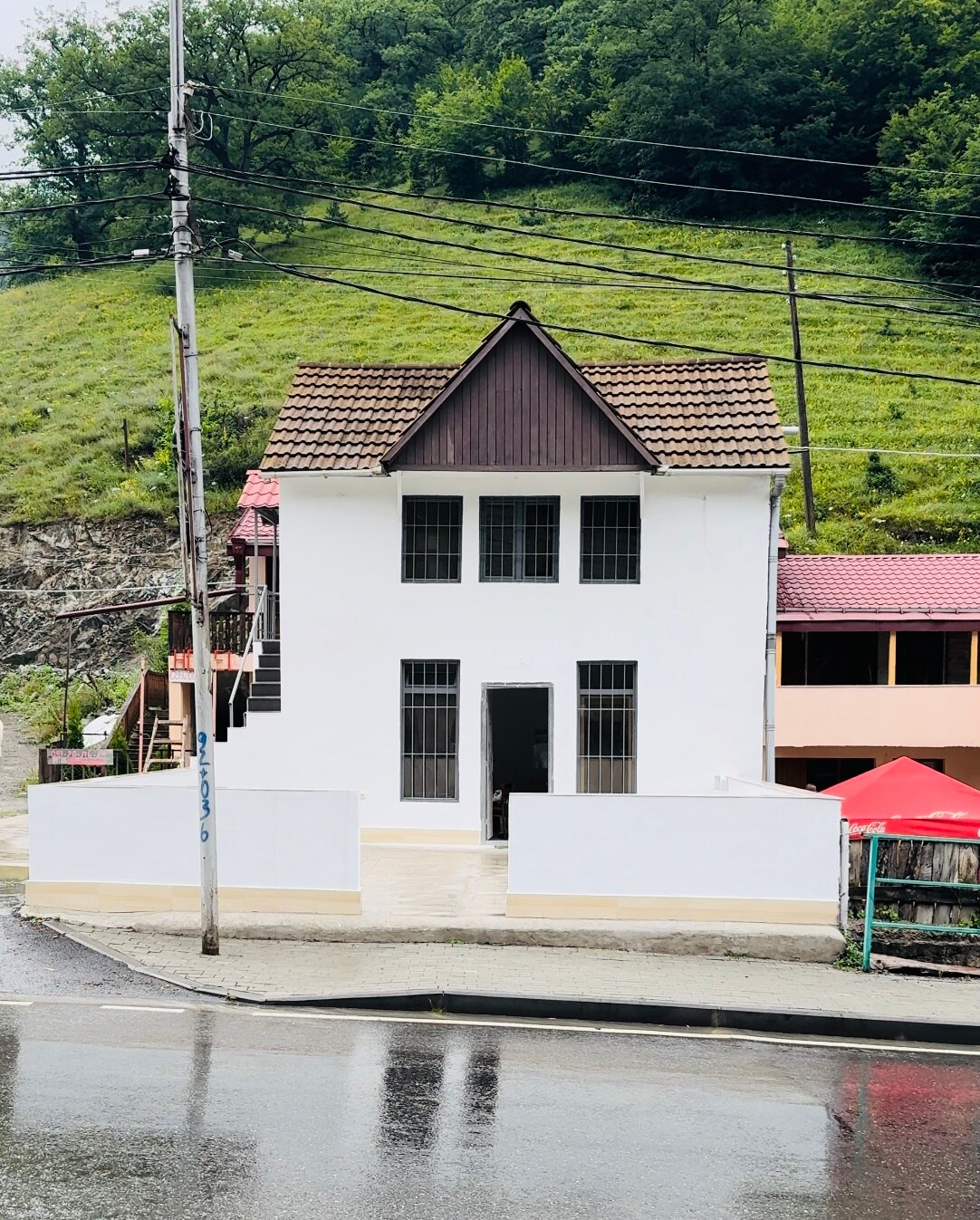 The exterior of a two-story house is shown, featuring white walls and a brown gabled roof. Large windows are present on both levels, allowing ample natural light. A porch area extends from the entrance, bordered by a low white wall. Green hills and other buildings are visible in the background.