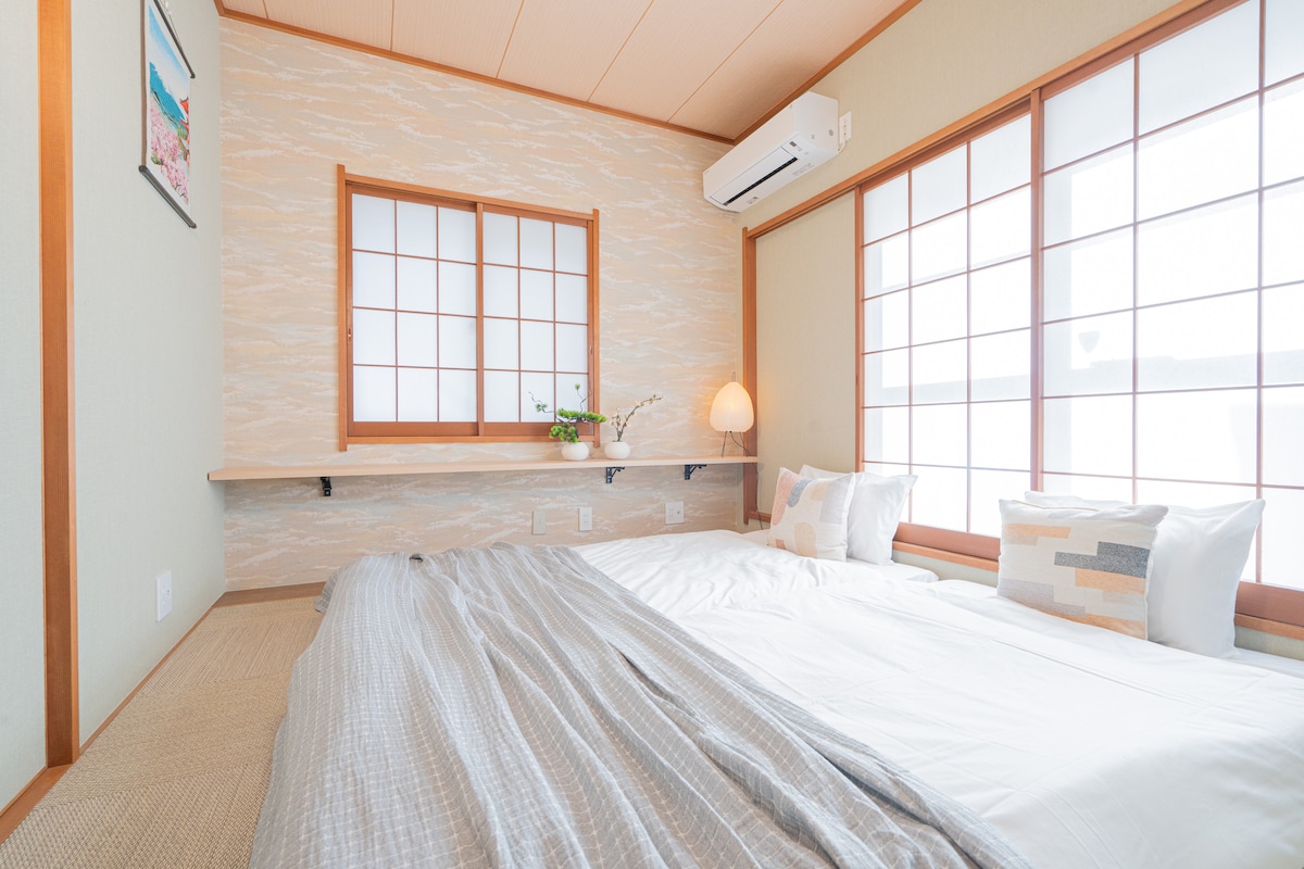 A traditional Japanese bedroom is highlighted with tatami mat flooring and soft lighting. Large windows with shoji screens allow natural light to fill the space. A spacious bed is dressed in neutral tones, complemented by minimalistic decor on the wall and a small plant on the shelf.