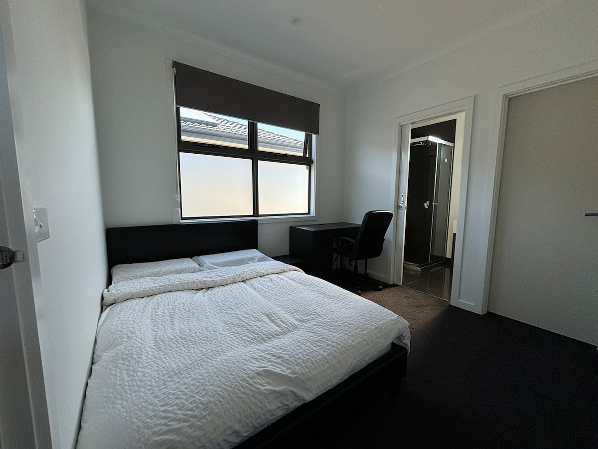 A well-lit master bedroom is depicted, featuring a spacious bed covered with a white duvet. A desk and chair are positioned nearby. An ensuite bathroom is visible through an open door, and a window allows natural light to enter the room.