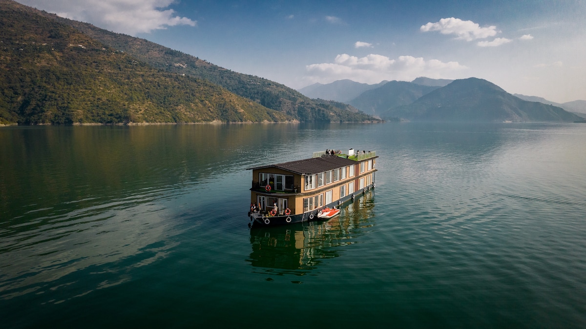 A modern houseboat is anchored on a calm lake, surrounded by lush, green hills and distant mountains. The structure features large windows and a rooftop area, providing an unobstructed view of the serene water and scenic landscape.