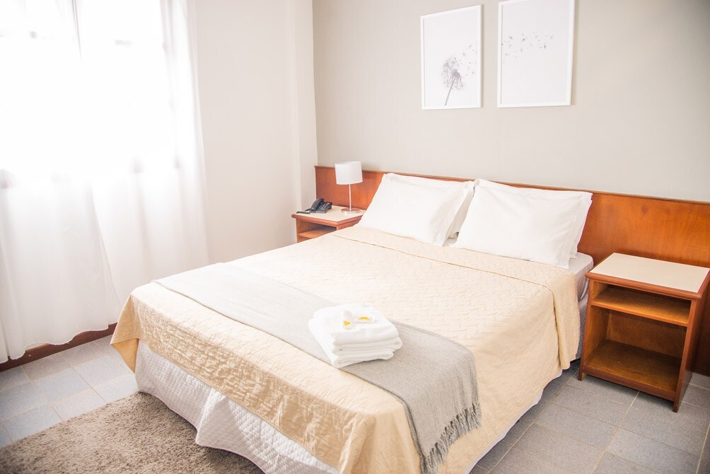 A cozy sleeping area features a spacious bed covered with a light beige blanket. Fluffy white towels are neatly arranged at the foot of the bed. Soft natural light filters through sheer curtains on a window, while simple wall art adds a touch of elegance.