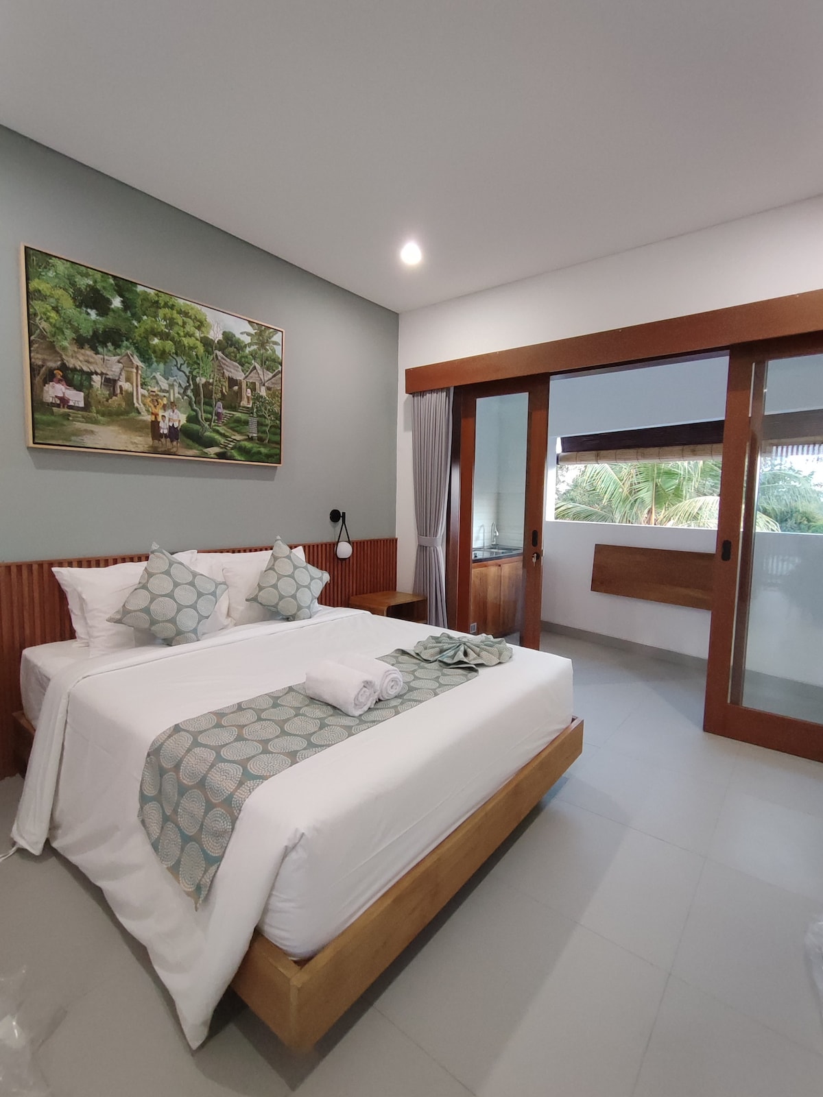 A modern bedroom features a spacious bed with decorative pillows and a light blanket. Artwork depicting a serene landscape adorns the wall. Natural light filters through large windows, offering views of greenery. A bedside lamp and a wooden table are also present.