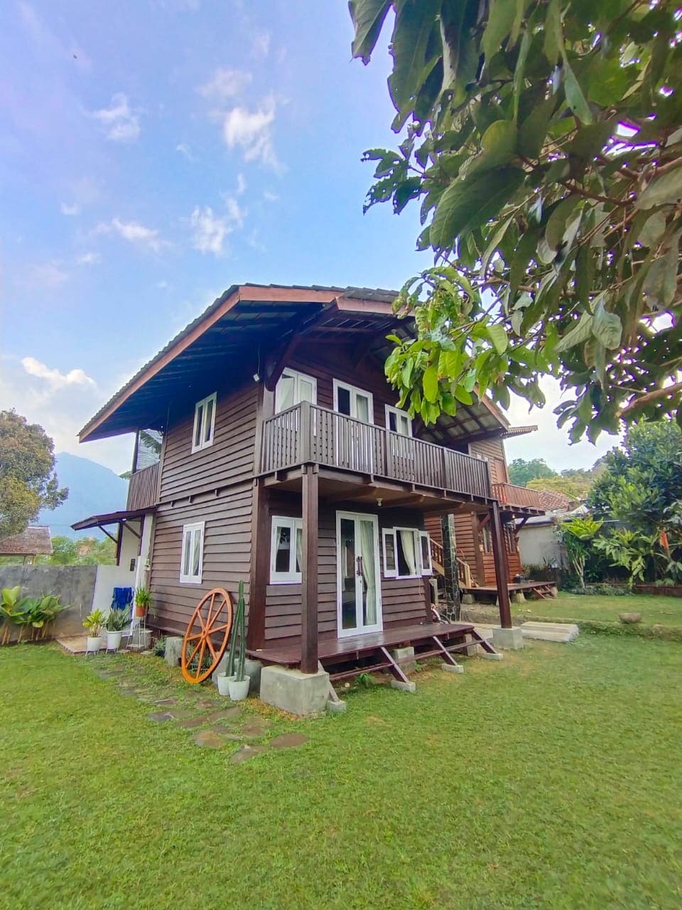 A two-story wooden house is situated in a lush green environment. The structure features a spacious porch with steps leading to the garden, framed by natural foliage. An inviting wheel decoration adds rustic charm to the exterior.