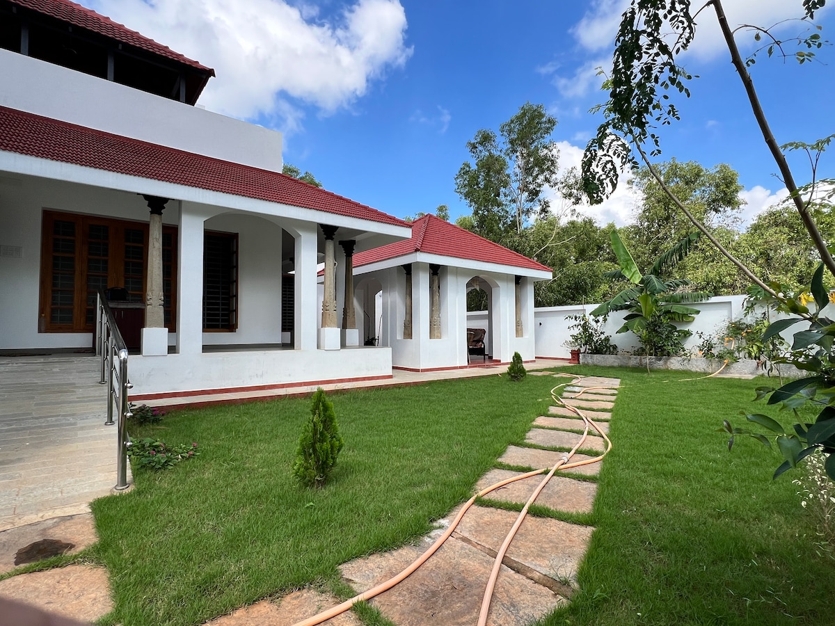 The exterior of the house is viewed, featuring a well-maintained lawn with stepping stones leading to a covered porch. Lush greenery surrounds the property, complemented by a clear blue sky and white clouds. A separate structure is visible at the back, enhancing the outdoor ambiance.