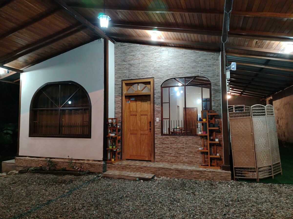 The exterior of a charming house is shown, featuring a wooden door framed by two arched windows. A stone-textured wall adds character, while soft lighting illuminates the entrance. A gravel path leads to the doorway, with a backdrop of lush greenery visible beyond a privacy screen.