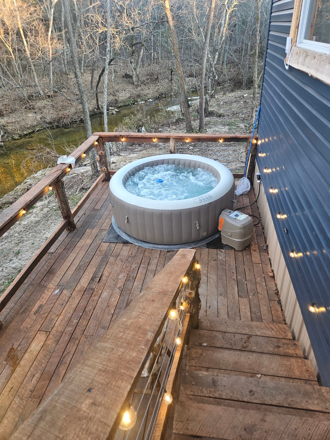 An inflatable hot tub is situated on a wooden deck, surrounded by a forest setting. Warm light strings are draped along the railing, illuminating the space. Below, a shallow creek is visible, bordered by trees that create a serene atmosphere.