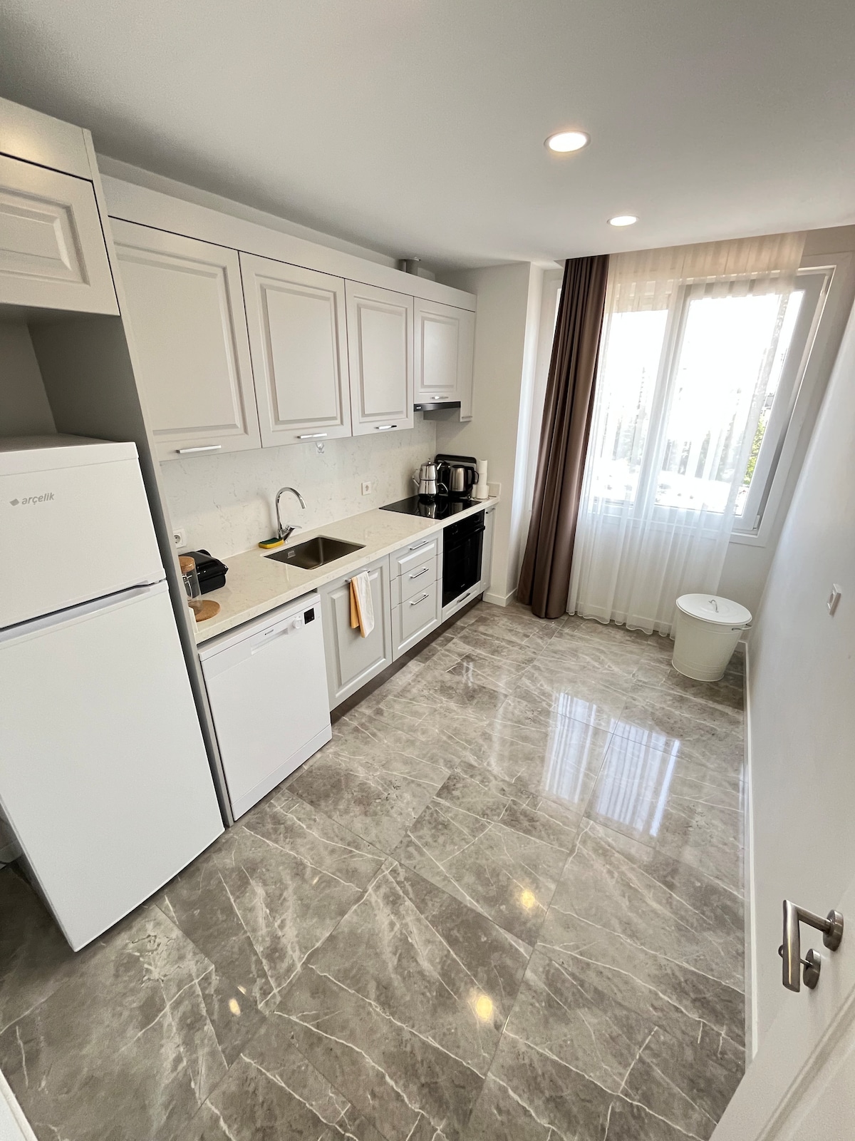 A modern kitchen is showcased with sleek cabinetry and a marble floor. White appliances and a black stove are present, along with a countertop that offers ample workspace. Natural light filters through a window adorned with sheer curtains, illuminating the space.