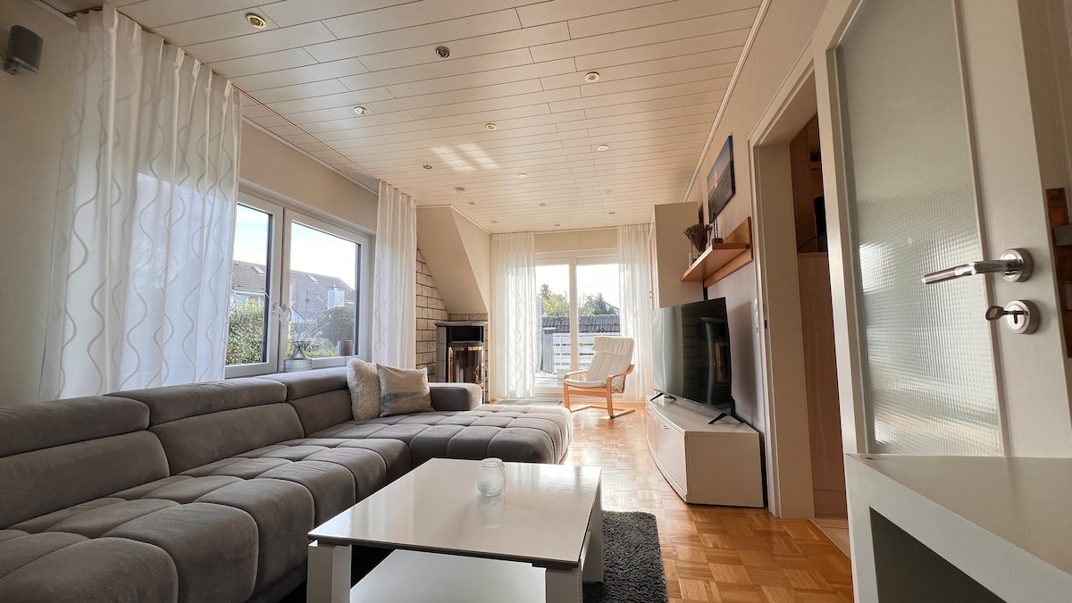A spacious living area is showcased, featuring a large grey sectional sofa and a white coffee table. Natural light floods through expansive windows, illuminating the room. A flat-screen television is mounted on the wall, with a cozy chair located nearby, creating a relaxed seating arrangement.