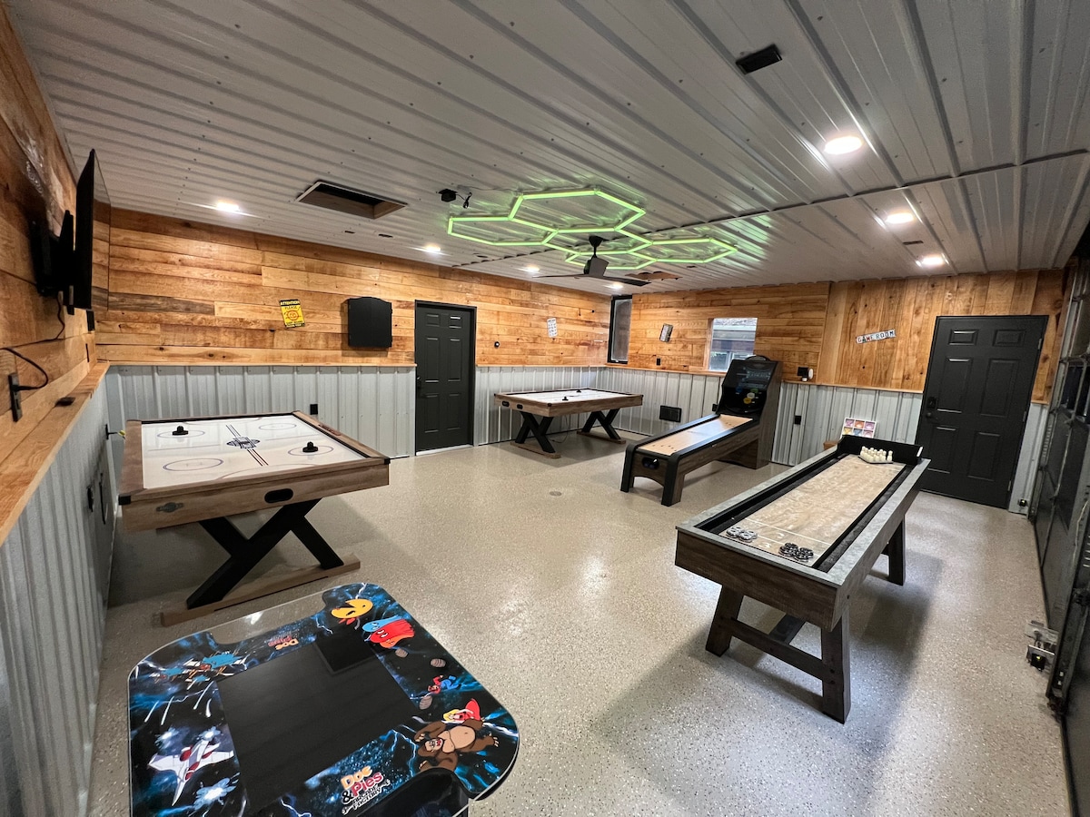 A spacious game room features a variety of entertainment options, including air hockey, shuffleboard, and a classic arcade game. The walls are adorned with wood paneling, and unique green LED lighting is positioned on the ceiling. Two solid tables are placed strategically across the room.