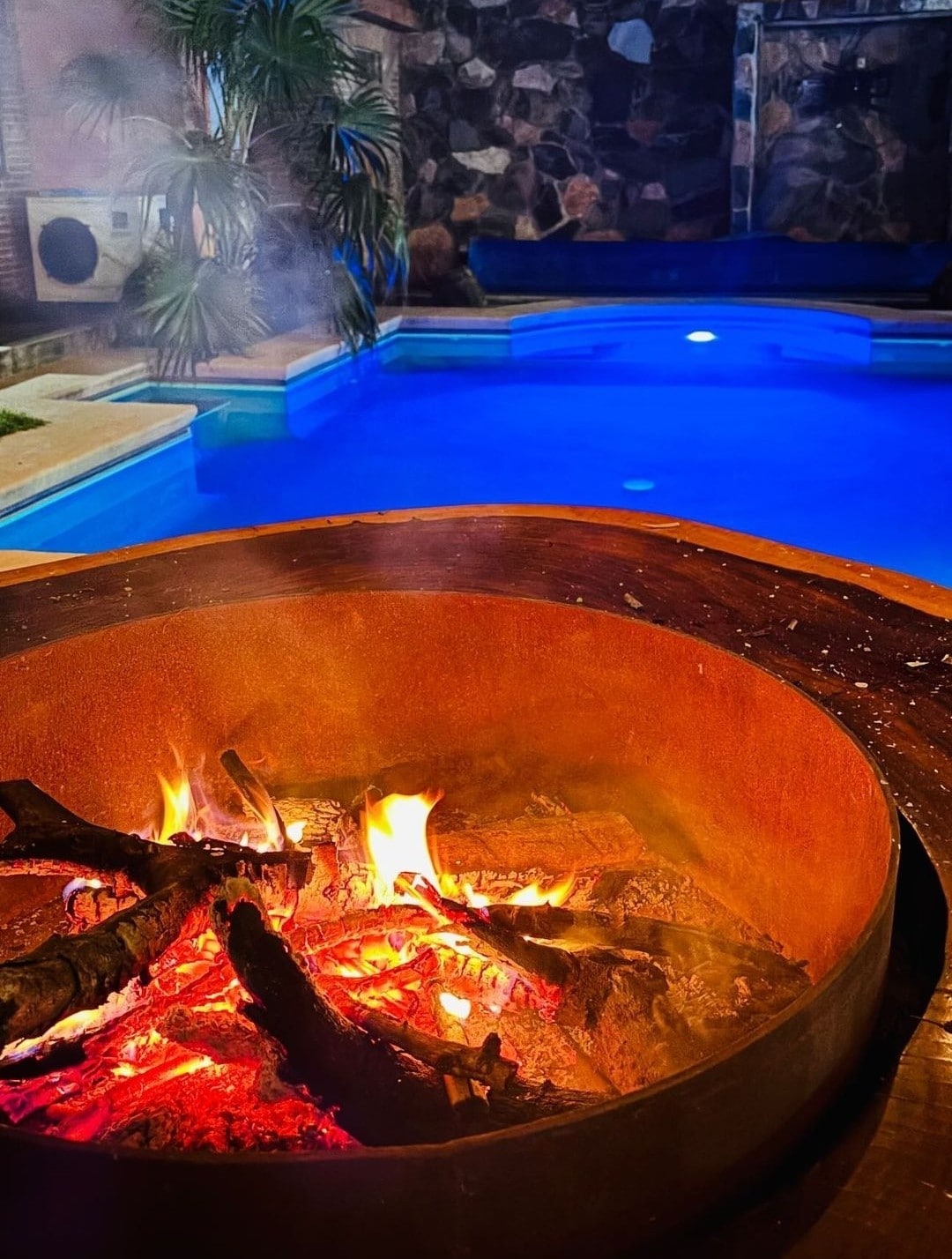 A circular fire pit sits in the foreground, with glowing embers and flames visible among the logs. In the background, a tranquil pool is highlighted in calming blue tones, surrounded by lush greenery and natural stone walls.